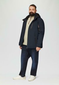 Marineblau isolierte Jacke mit Kapuze, Reißverschlusstaschen und kontrastierenden Reißverschlüssen, getragen über einem hellbeigen Pullover und dunklen Jeans.