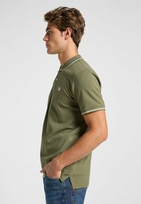 Oliivivihreä polo-paita, jossa on lyhyet hihat, kaksi nappia ja kontrastivalkoiset kauluskoristeet. Valmistettu pehmeästä kankaasta, jossa on sivuhalkiot.