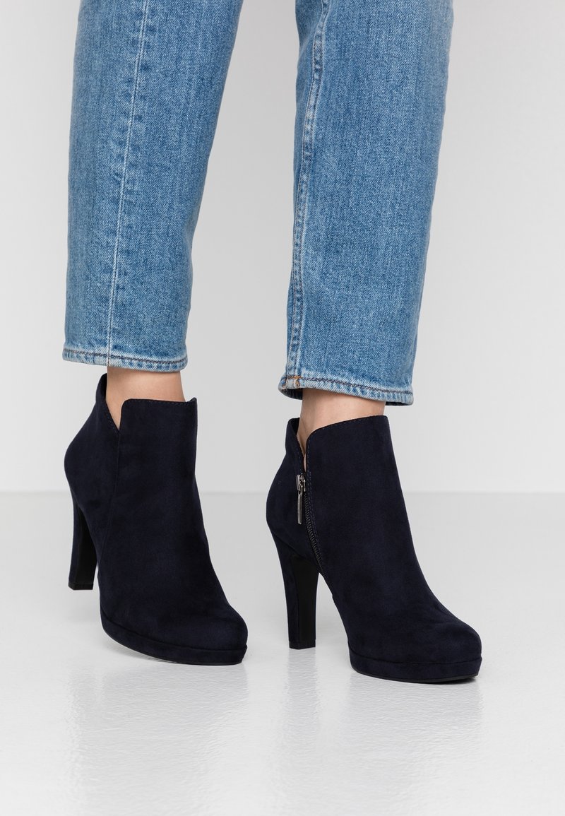 Personne portant un jean en denim bleu et des bottines noires en daim à talons hauts avec des fermetures éclair latérales, debout sur un sol blanc brillant.