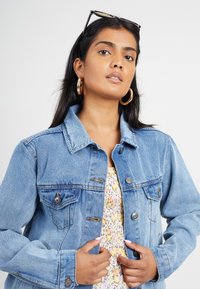 Giacca di jeans azzurro chiaro con due tasche sul petto, chiusura con bottoni e colletto classico. Texture liscia con zone leggermente sbiadite.