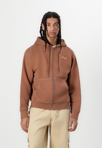 Brown zip-up hoodie with front pockets, drawstring hood, contrasting stitching, and a small yellow logo. Worn with light-colored pants.