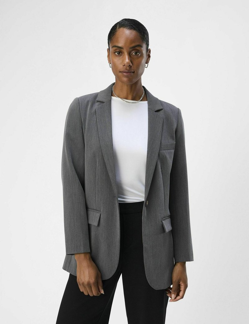 Blazer ajusté gris avec revers crantés, fermeture à un bouton et deux poches frontales. Associé à un haut blanc et un pantalon noir.