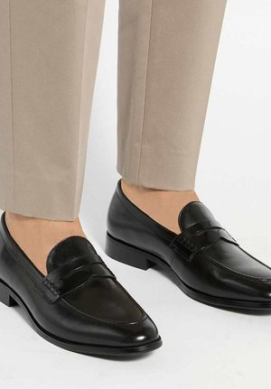 Mocassins penny en cuir noir portés avec un pantalon beige court, présentés aux pieds sur un fond blanc.