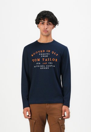 Junger Mann mit dunklem, lockigem Haar, trägt ein marineblaues Langarmshirt mit orangefarbener und weißer Schrift, kombiniert mit braunen Cargo-Hosen, steht vor einem weißen Hintergrund.