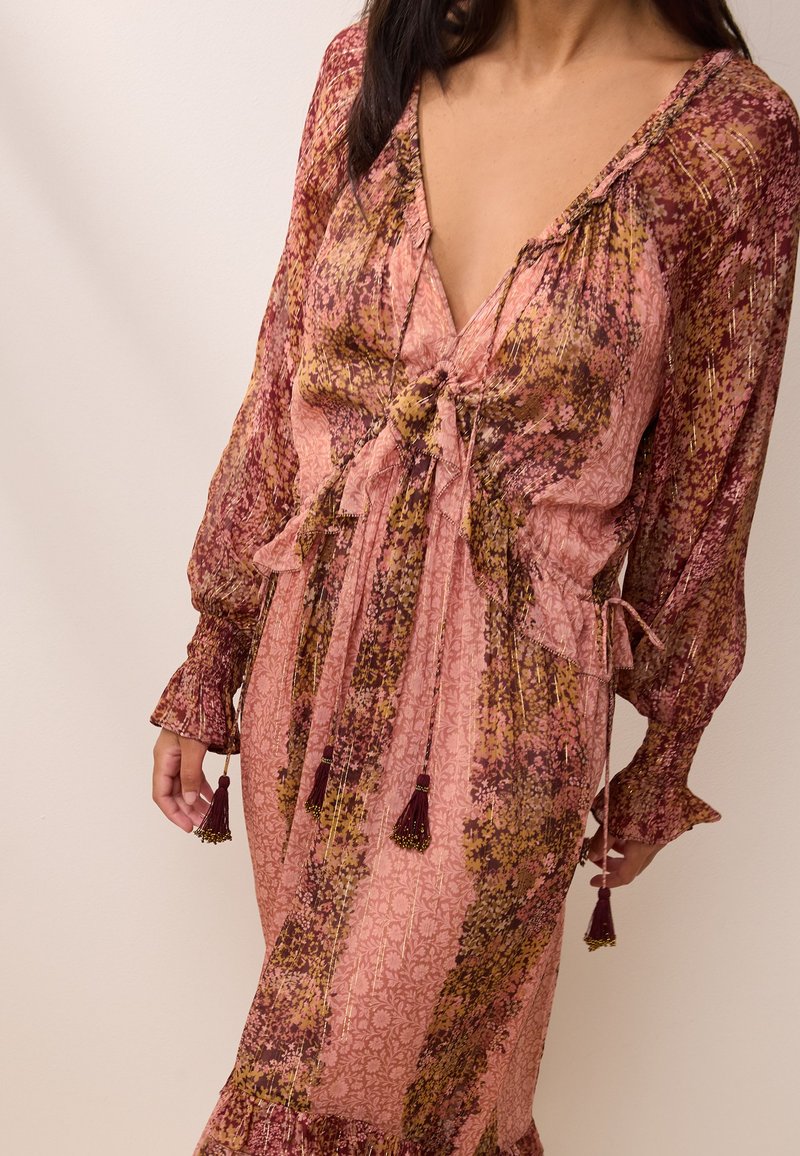 Femme portant une robe à manches longues rose et bordeaux à motif avec des détails à nouer et des pompons au niveau de l'encolure et des manches.