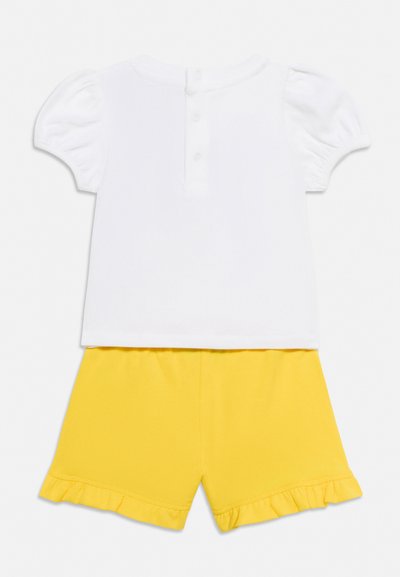 Chemise blanche à manches courtes avec boutons, associée à un short jaune avec ourlets à volants, présentée à plat sur un fond uni.