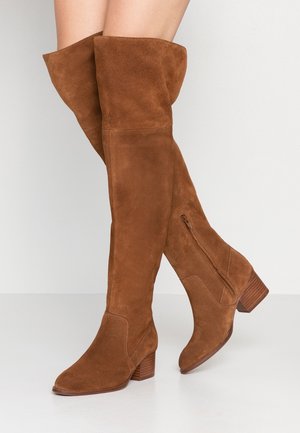 Bottes montantes en daim marron avec talons blocs empilés, fermeture éclair latérale et bouts légèrement froncés, portées sur des jambes croisées.