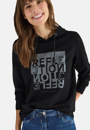 Ung kvinde, der smiler, iført en sort hoodie med en grå reflekterende firkant og teksten "REFLECTION" og "find din indre lys", kombineret med blå jeans.