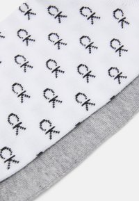 Calvin Klein čarape u bijeloj i sivoj boji. Bijela čarapa ima ponovljeni crni „CK” logo; siva čarapa ima glatku teksturu.