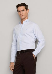 Muž v svetlomodrej pruhovanej košeli na gombíky a tmavohnedých nohaviciach s rukou v vrecku, stojaci pred jednoduchou béžovou stenou.