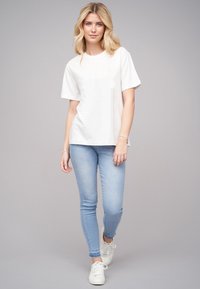 Bílý tričko s krátkým rukávem v kombinaci s světle modrými skinny džínami a bílými teniskami, na neutrálním pozadí.
