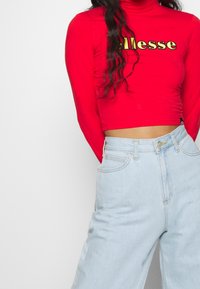 Roter Langarm-Cropped-Top mit goldfarbenem "ellesse"-Logo, kombiniert mit hellblauen weiter geschnittenen Jeans mit Fronttaschen und hohem Bund.