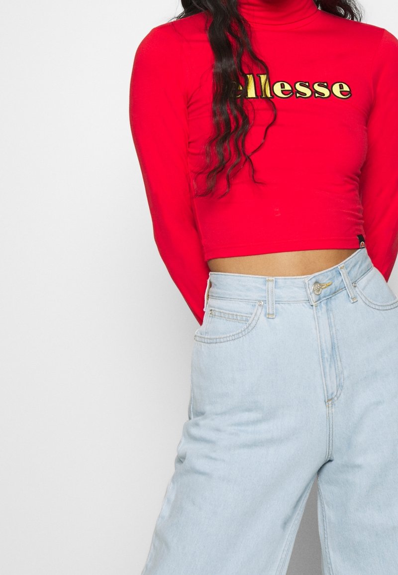 Roter Langarm-Cropped-Top mit goldfarbenem "ellesse"-Logo, kombiniert mit hellblauen weiter geschnittenen Jeans mit Fronttaschen und hohem Bund.