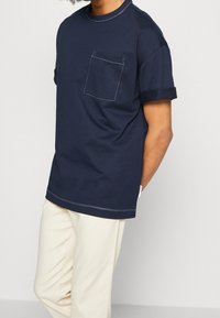 Granatowy bawełniany t-shirt z krótkimi rękawami, podwiniętymi mankietami, kieszenią z przodu oraz kontrastującymi białymi przeszyciami na dekolcie i dole.