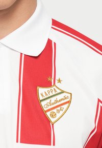 Polo tričko s červenými a bielymi pruhmi a golierom. Obsahuje vyšitú zlatú nášivku s „Kappa Authentic 1967“ a dvoma hviezdami.