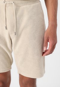 Pantalones cortos de rizo beige con cintura ajustable, que cuentan con una textura suave y un corte relajado. Acentos de metal en plata visibles.
