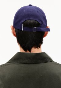 Marineblaues Baseballcap mit verstellbarem Riemen auf der Rückseite, getragen über dunklem Haar, kombiniert mit einem dunkelgrünen Hemd mit Kragen.