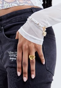 Gold heart-shaped ring with "KISSES" engraving, worn on a finger. White sheer long-sleeve top and pearl-accented bracelet visible. Dark jeans.