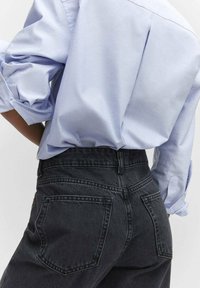 Vaqueros negros de cintura alta con un diseño clásico de cinco bolsillos, combinados con una camisa azul claro abotonada con las mangas remangadas.