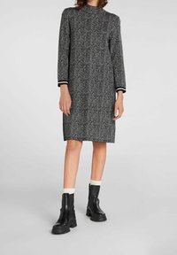 Robe à hauteur de genoux en pied-de-poule noir et blanc, avec des manches longues et un col haut, présentant des poignets rayés noir et blanc. Assortie à des bottes noires épaisses.