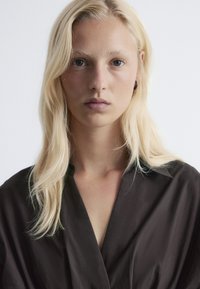 Model with medium-length straight blonde hair, wearing a dark brown collared shirt. Soft texture, minimal detailing, neutral background.