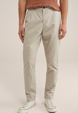 Pantalones de algodón beige con corte recto, pliegues frontales y cierre de botón. Combinados con zapatillas blancas de perfil bajo.