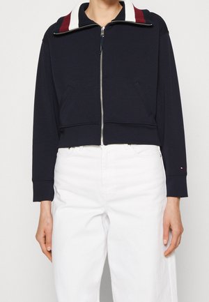 Personne portant une veste à fermeture éclair bleu marine avec des rayures rouges et blanches sur le col et un pantalon blanc taille haute, debout devant un fond uni.