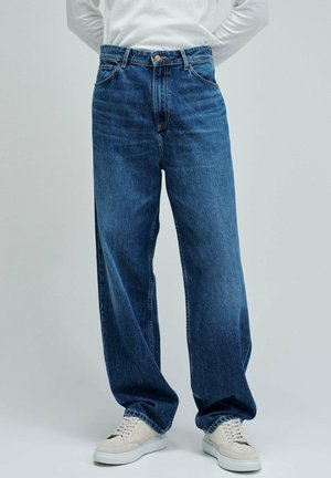 Personne portant un jean bleu ample et des baskets blanches, debout devant un fond gris clair uni.