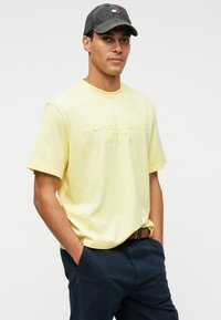 Geltonas trumpas rankovės marškinėlis su toniniu "Tommy Jeans" logo. Derinkite su tamsiomis kelnėmis ir tamsia kepure, kad sukurtumėte kasdienį įvaizdį.