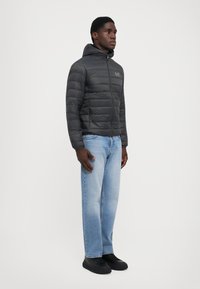 Veste noire rembourrée avec une fermeture éclair et une capuche, présentant un design matelassé, portée avec un jean bleu clair et des chaussures noires.