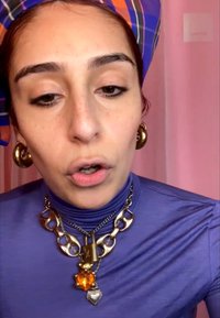 Femme aux cheveux roux portant un col roulé bleu, un collier chaîne épais en or avec des breloques cadenas et cœur, des boucles d'oreilles créoles dorées assorties et un foulard à motifs.