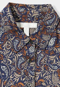Bluse mit Blumenmuster in navy-basis, mit orange und cremefarbenen Designs. Hemdkragen mit Knopfleiste und zwei sichtbaren Knöpfen. Der Stoff hat eine Textur.