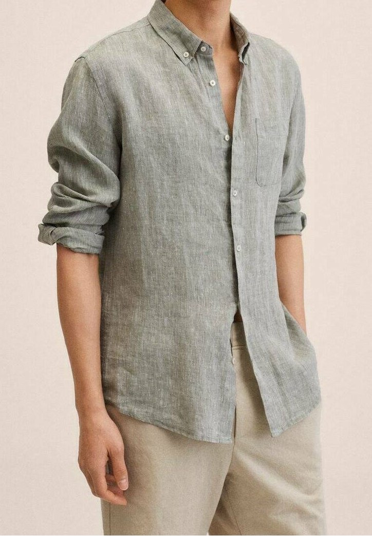 Homme portant une chemise en lin gris clair à manches longues retroussées et un pantalon beige, debout avec une main dans la poche.