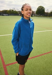 Blå fleecejakke med lynlås foran, sidelommer og høj krave, kombineret med sorte shorts på en grøn sportsbane.