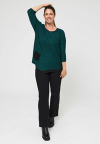 Turkizna teksturirana majica s crnim džepom, rukavi do tri četvrtine i zakrivljenim rubom, uparena s crnim hlačama i čizmama do gležnja.