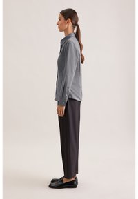 Camicia grigia con bottoni, colletto e maniche risvoltate, abbinata a pantaloni neri su misura e mocassini neri lucidi, vista di lato.