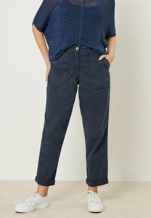 Persona con pantalones azul marino con puños y grandes bolsillos delanteros, zapatillas blancas y un suéter de punto azul marino holgado, de pie con las manos en los bolsillos.