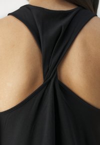 Lähikuva henkilön yläselästä, joka pitää yllään mustaa hihattomaa toppia, jossa on kierrerakenteinen kangassuunnittelu keskellä.