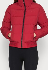 Czerwona kurtka puchowa z czarnym zamkiem, elastycznymi mankietami i logo Calvin Klein na dolnej części, noszona z czarnymi spodniami.