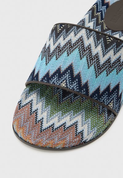 Sandale slide à motif zigzag coloré avec dessus en maille bleu, marron, vert et noir, bordure noire sur fond clair.