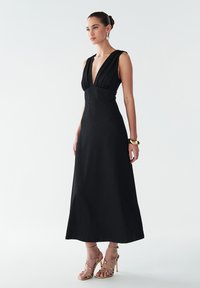 Robe maxi noire avec un décolleté en V profond, tissu texturé, corsage ajusté et jupe fluide. Associée à des talons dorés à lanières et à un bracelet.