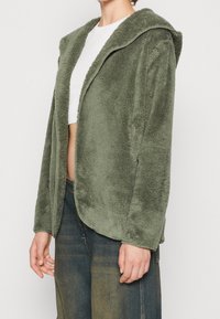 Grön fleecejacka med huva, med en mjuk textur och öppen framsida. Matchad med lösa denimbyxor.