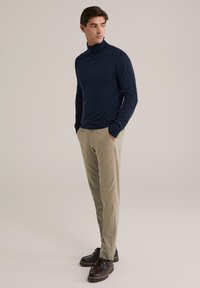 Hombre vistiendo un suéter de cuello alto azul marino, pantalones beige y zapatos de cuero marrón, de pie con las manos en los bolsillos, mirando de lado.