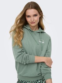 Jeune femme aux longs cheveux blonds, portant un sweat à capuche vert sauge avec le texte "LIMITLESS" et des leggings verts à motifs, posant devant un fond uni.