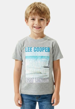 Jeune garçon souriant portant un t-shirt gris Lee Cooper avec un texte bleu et une image de voilier, assorti à un jean bleu, sur fond blanc.