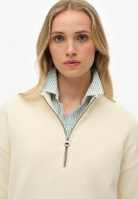 Off-white zip-up sweater met een geribbelde kraag, gedragen over een lichtblauw en wit gestreept shirt, met een zichtbare zilveren rits.