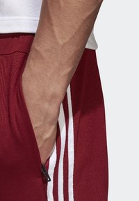 Red athletic shorts with white stripes, featuring a side zipper pocket. The waistband is elastic; the fabric has a smooth texture.