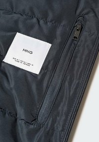 Tissu matelassé noir avec une fermeture éclair verticale noire et une étiquette blanche indiquant la marque MNG et la taille moyenne pour EUR, USA et MEX.