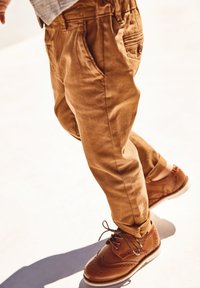 Tan cotton trousers with pockets, paired with brown leather shoes featuring perforated accents and laces. Designed for casual wear.