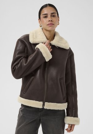 Frau trägt dunkelbraune Lederjacke mit cremefarbenem Lammfellkragen, Manschetten und Saum, steht vor hellem, einfarbigem Hintergrund.